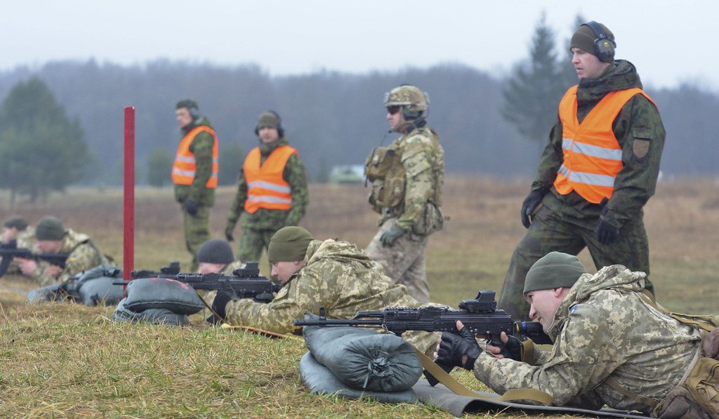 A live fire line with prone shooters and safety staff in high visibility vests standing behind them