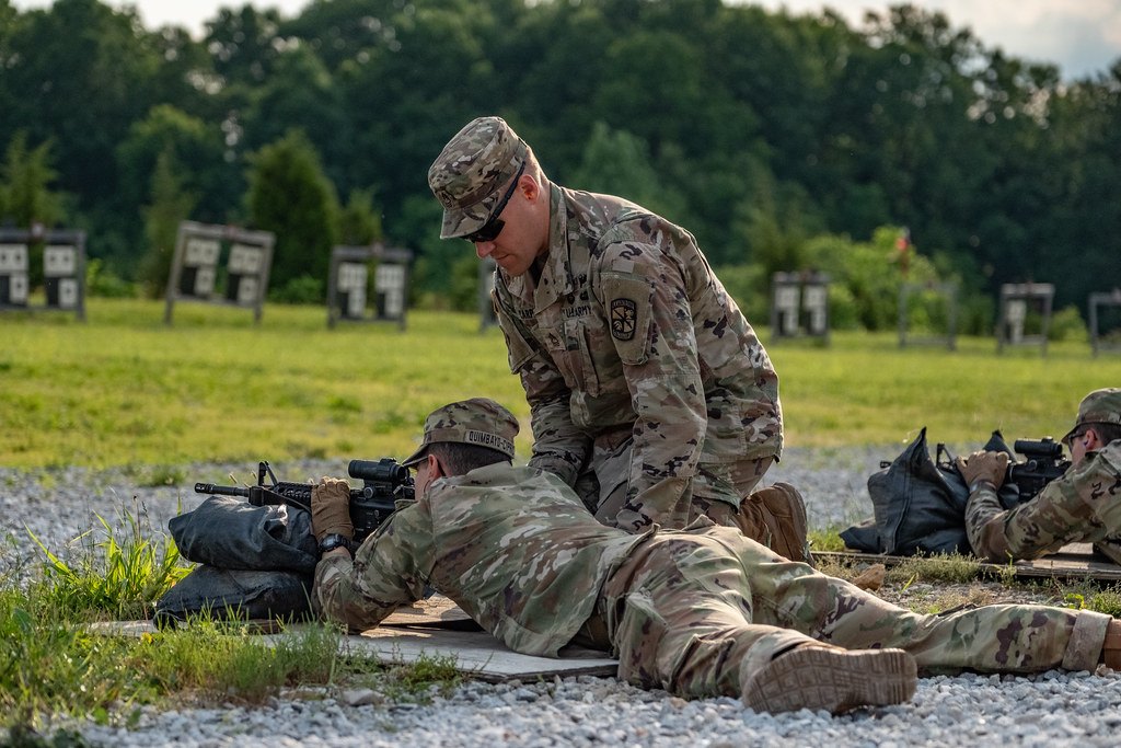 An instructor kneeling beside a prone shooter on a gravel firing line, with target frames in the distance