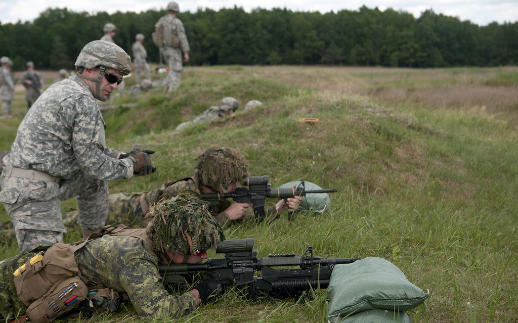 An instructor kneeling beside shooters lying prone in long grass on a range