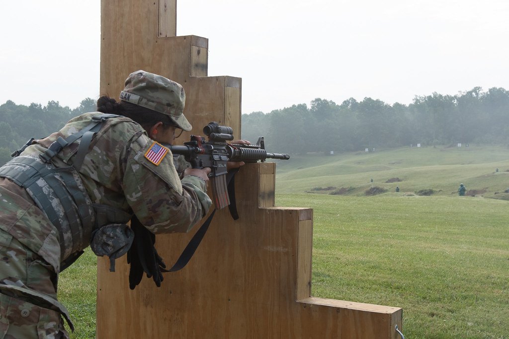 A shooter leaning around the left side of a wooden barricade with a rifle up, on a misty range