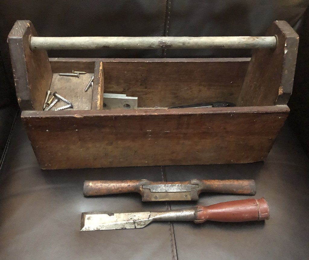 An old wooden tool tote holding screws and hardware, with two chisels laid in front of it