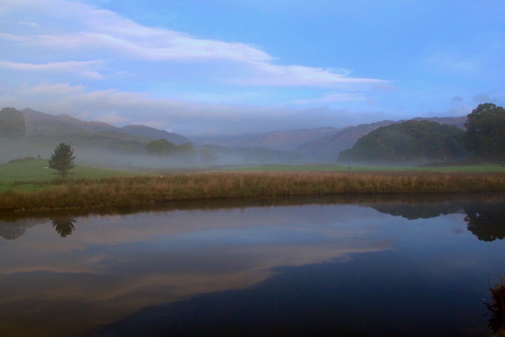 Mist lying across water and low hills at first light, with rough grass along the near bank