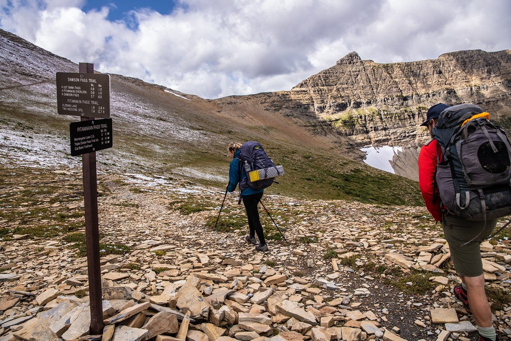 Three people carrying large backpacks walking across a boulder field below a snow streaked mountain range