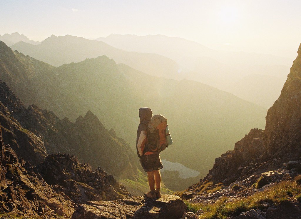 A hiker wearing a backpack standing on a rocky ridge above hazy mountain ridgelines at sunrise