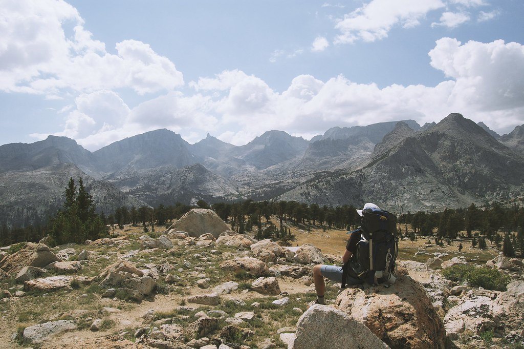A hiker with a large backpack sitting on a boulder looking towards a granite mountain range