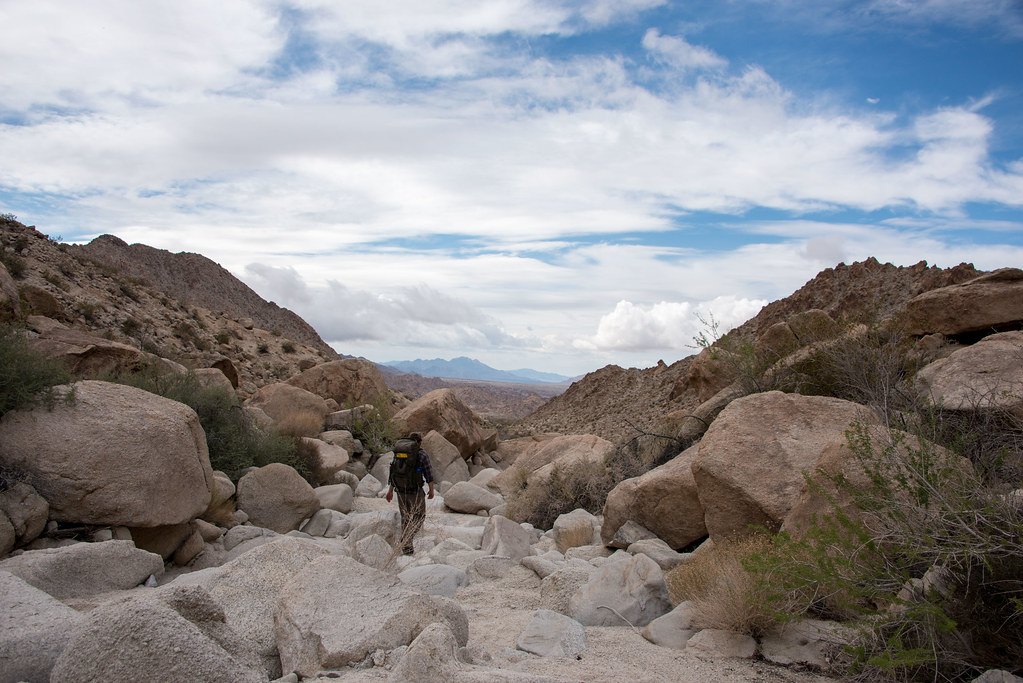 A person with a backpack walking up a boulder strewn wash between rocky desert hills