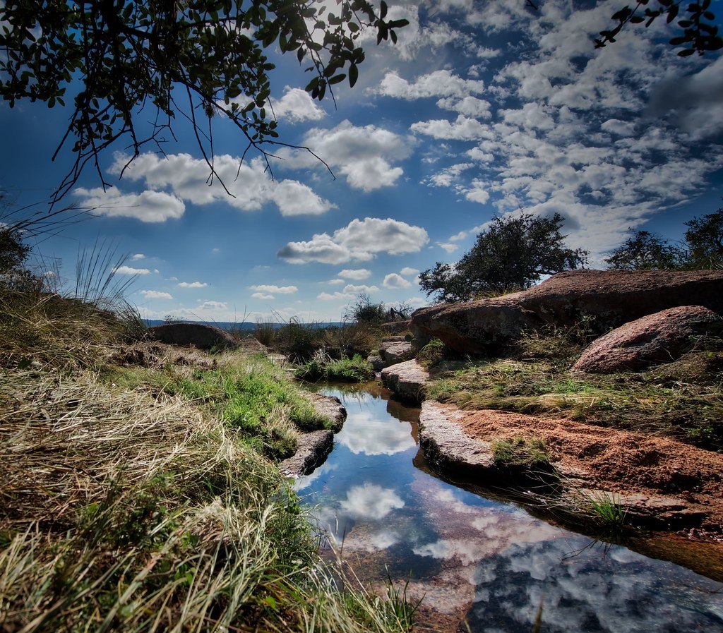 A shallow stream running over pink granite among live oaks and grass under a cloudy sky