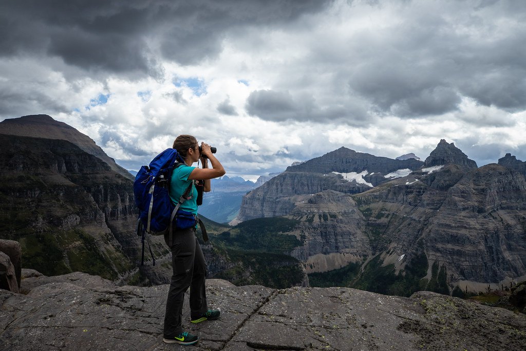 A person wearing a backpack standing on a rock ledge looking through binoculars at a mountain valley