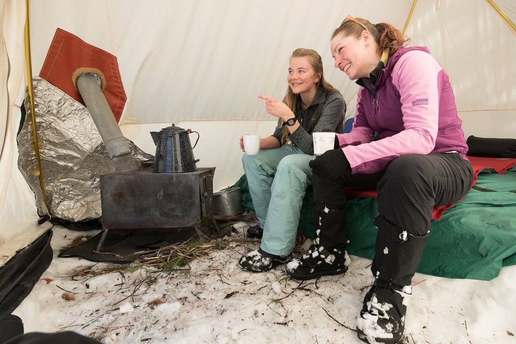 Two people sitting in a canvas wall tent beside a wood stove with snow on the ground