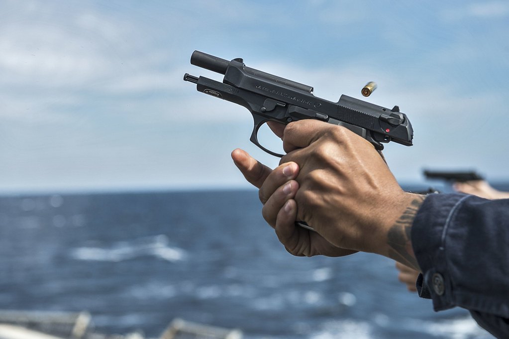 Close view of two hands wrapped around the grip of a semi-automatic pistol, the slide to the rear and a brass case in the air