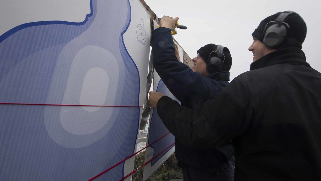 Two people wearing ear protection fixing a large paper silhouette target to a wooden frame on an outdoor range