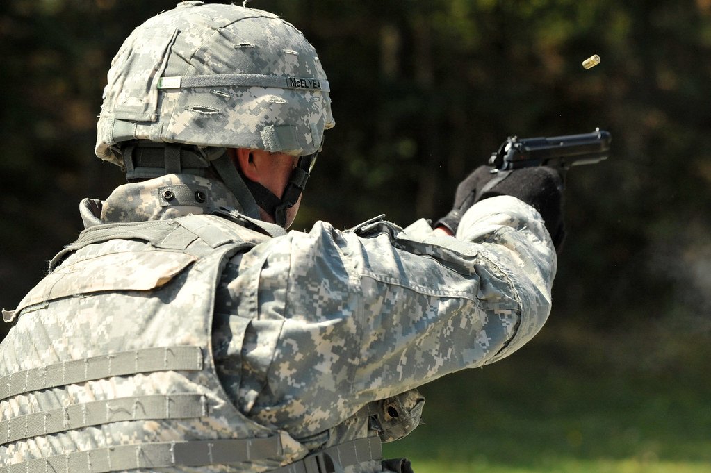A shooter seen from behind firing a semi-automatic pistol with both arms extended, a spent brass case in the air beside the slide