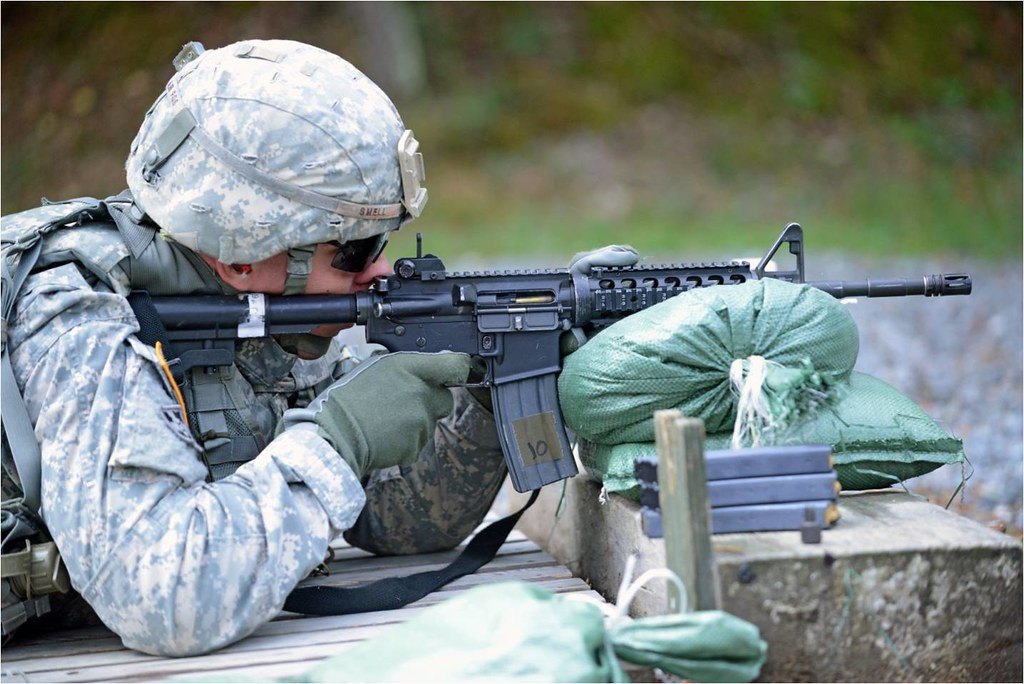A shooter lying prone behind a rifle rested on sandbags, looking through the sights on an outdoor range