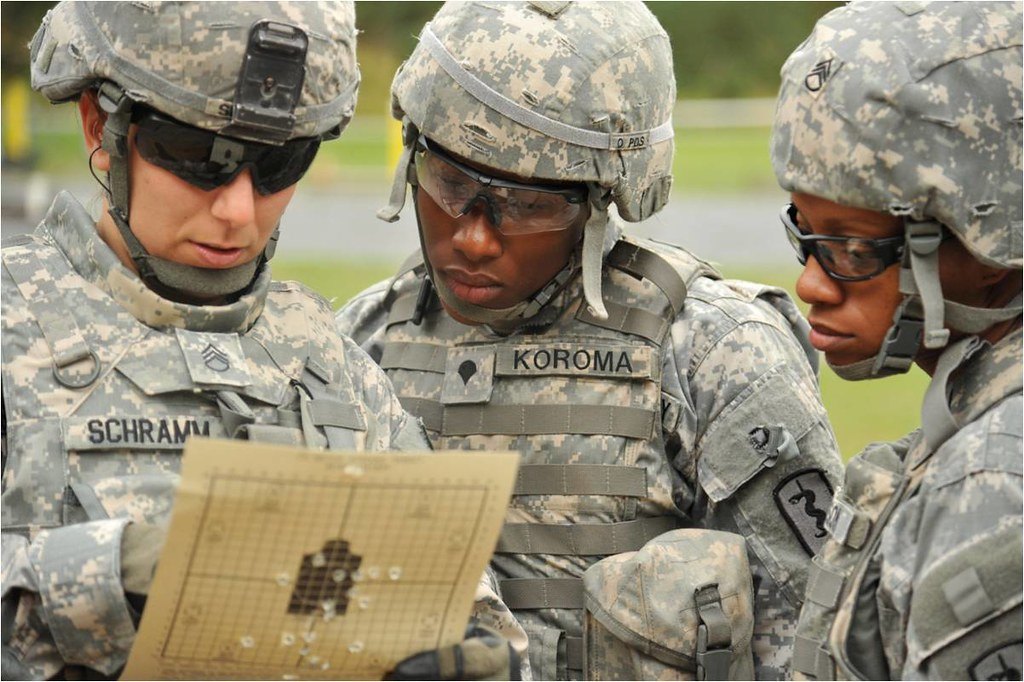 Three people in helmets and eye protection studying a paper target held between them, a cluster of bullet holes near its centre