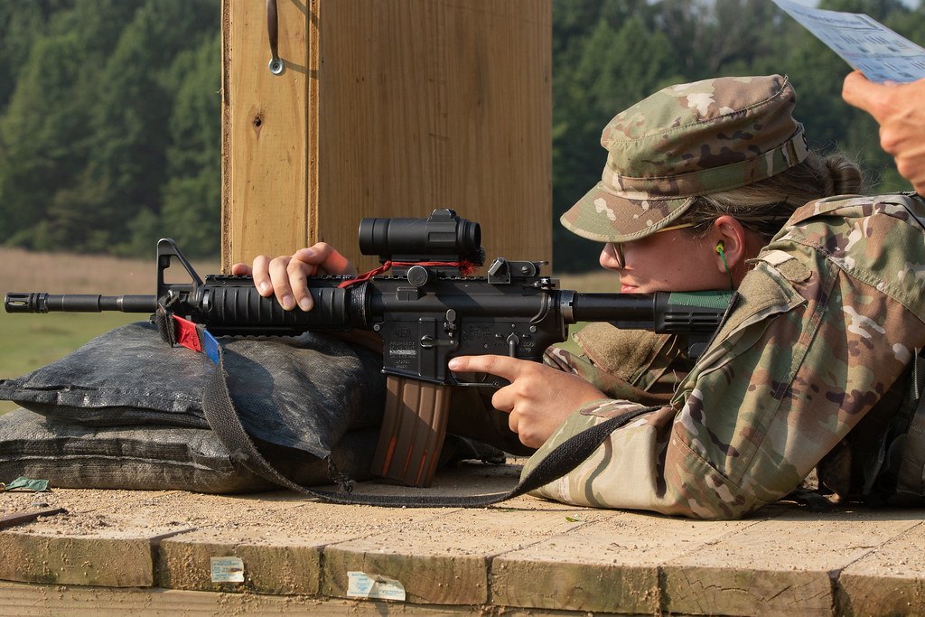 A shooter lying prone behind a rifle rested on sandbags while a second person beside them holds a scorecard