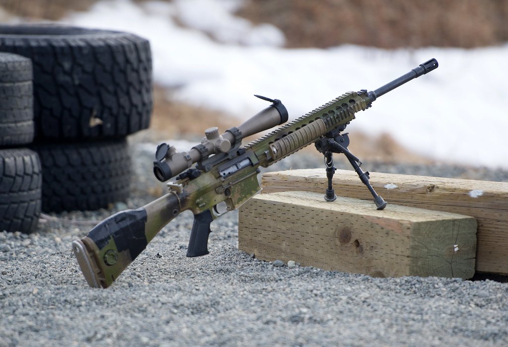 A rifle fitted with a telescopic sight and a folded bipod resting against a wooden beam on a gravel range, stacked tyres behind it