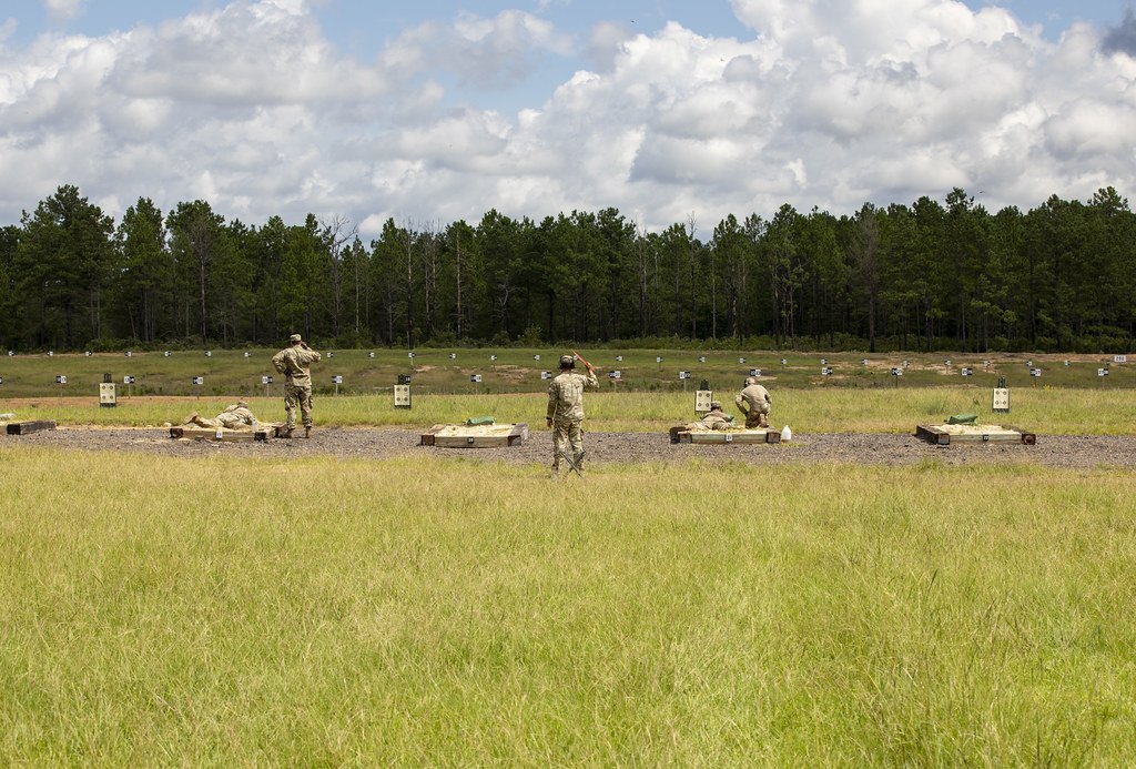 A wide grassy rifle range with shooters lying prone on firing points and paper target frames spaced along a berm in the distance
