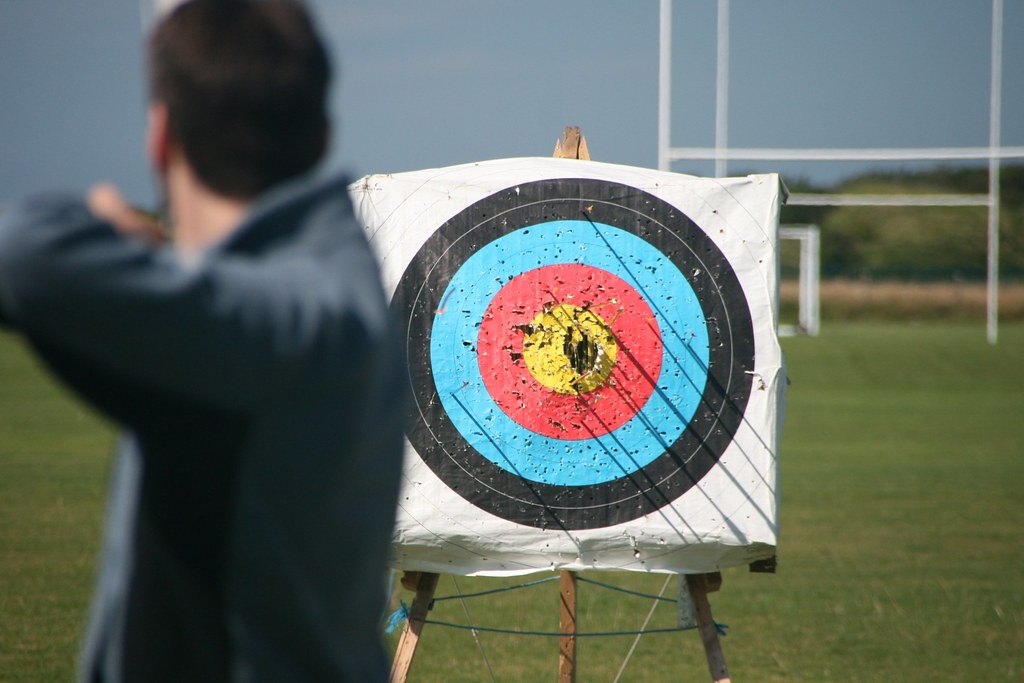 A round archery target face on a straw butt in a field, with an out of focus archer at draw in the foreground