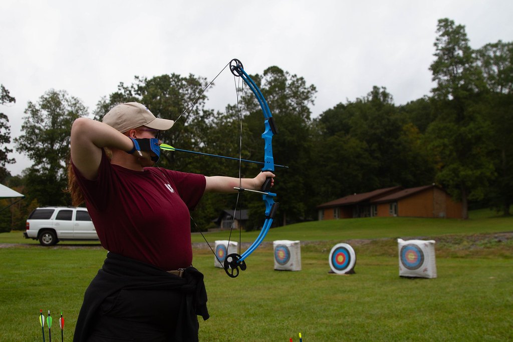 An archer at full draw with a blue compound bow on a grass field, several round target butts standing at different distances behind