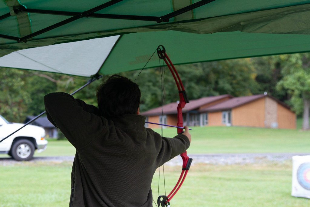 An archer at full draw with a red compound bow, shooting from under an open canopy toward a target butt on a lawn