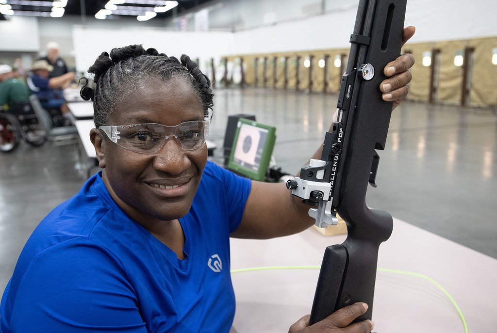 A person at an indoor airgun range holding a target air rifle upright, an electronic scoring monitor on the bench behind