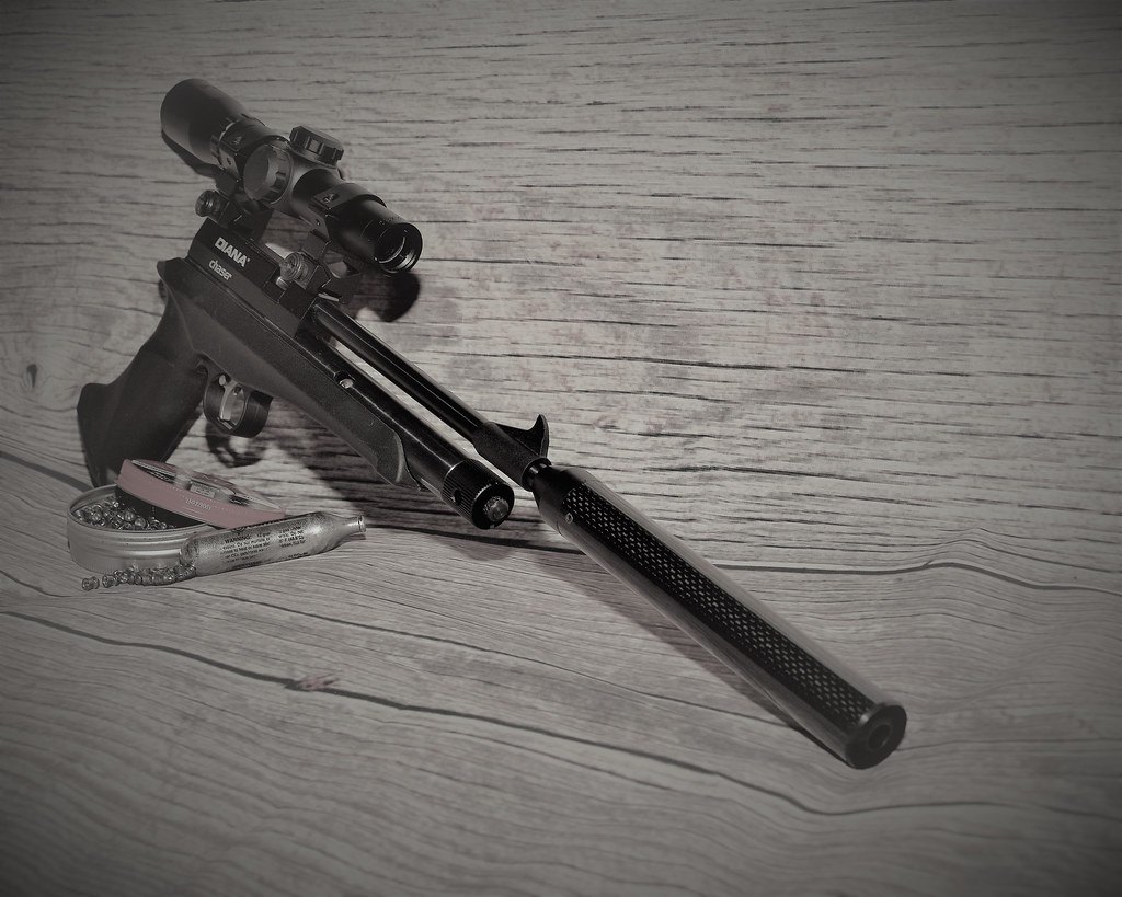 An airgun fitted with a telescopic sight lying on weathered wood beside an open tin of pellets and a gas cartridge
