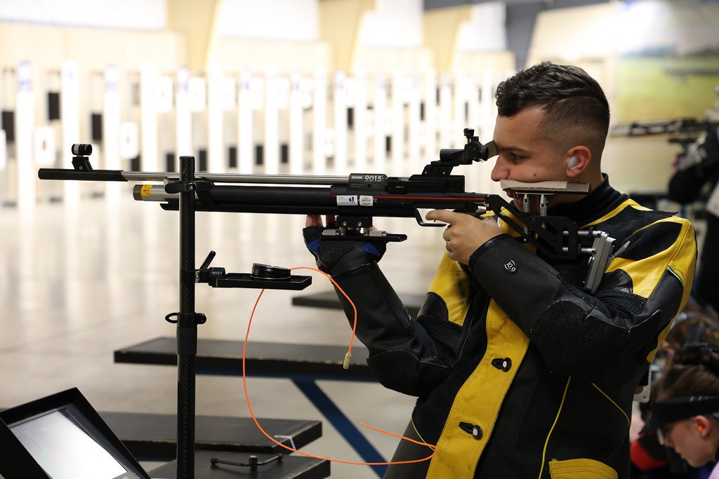 An air rifle shooter in a heavy shooting jacket standing at an indoor range, aiming at a row of paper targets down the lane