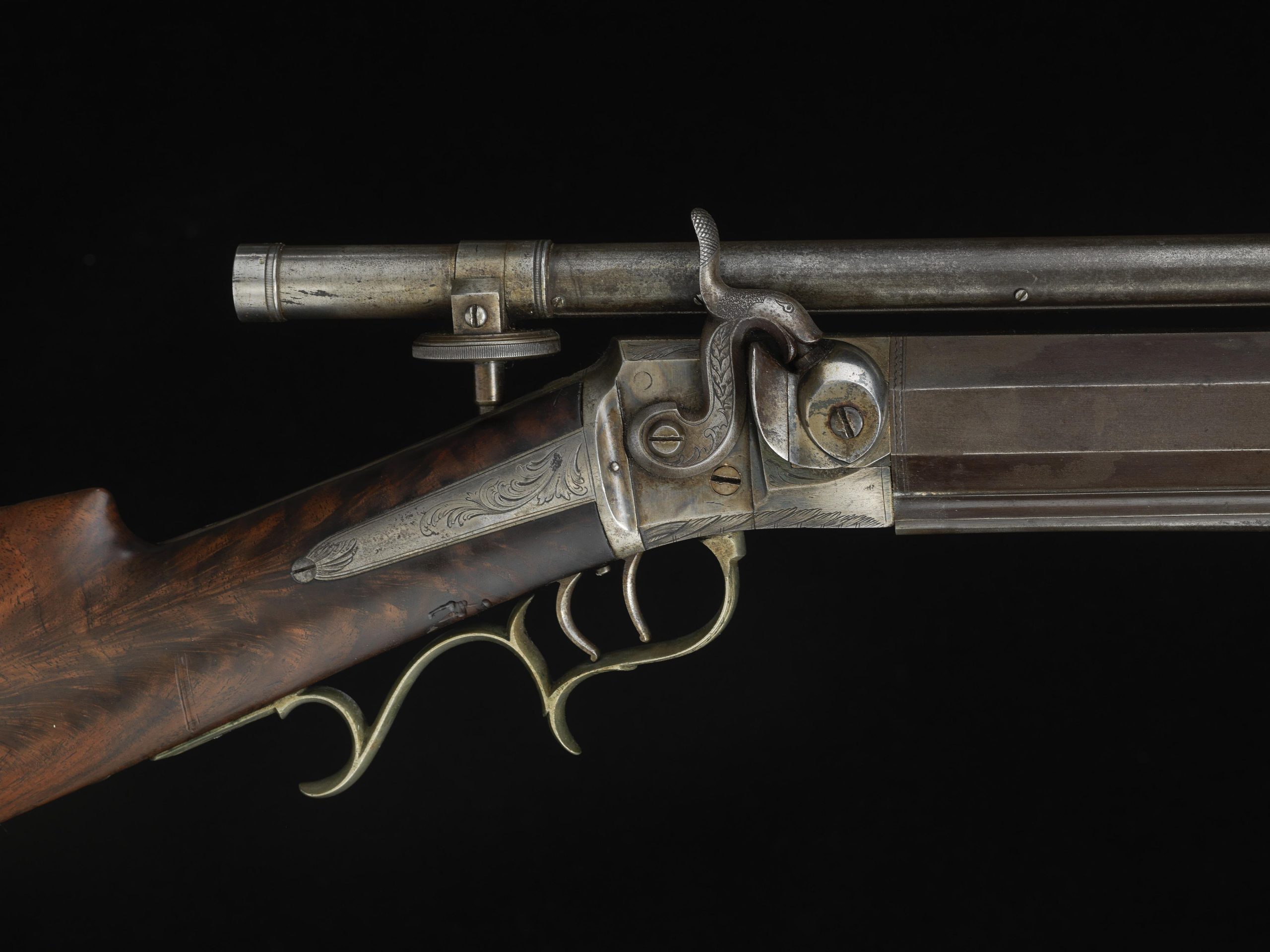 A muzzleloading match rifle with a long tube telescopic sight mounted along the top of the barrel, against a black backdrop