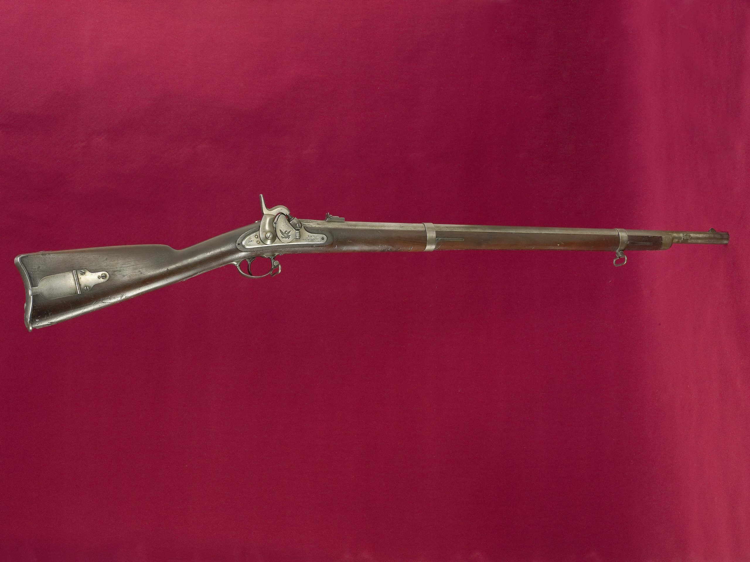 A percussion muzzleloading rifle with a wooden stock and long steel barrel, shown side on against a deep red backdrop