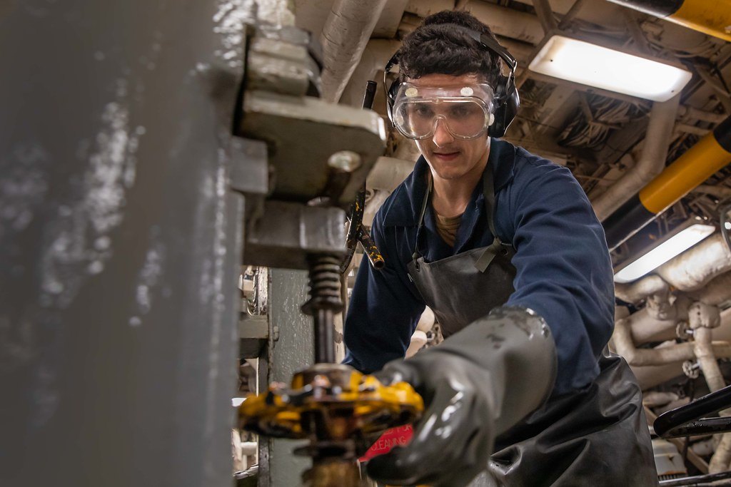 A worker in safety goggles and heavy gloves servicing machinery in an engine space
