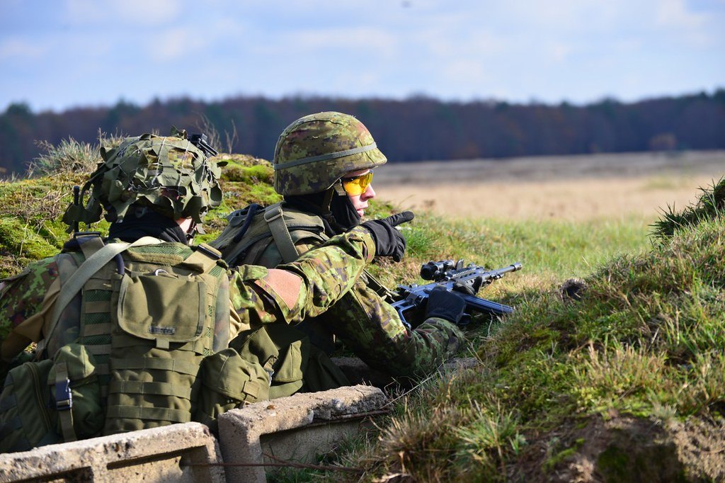 Two soldiers in a shallow field position behind a low concrete parapet, one with a rifle resting on the edge